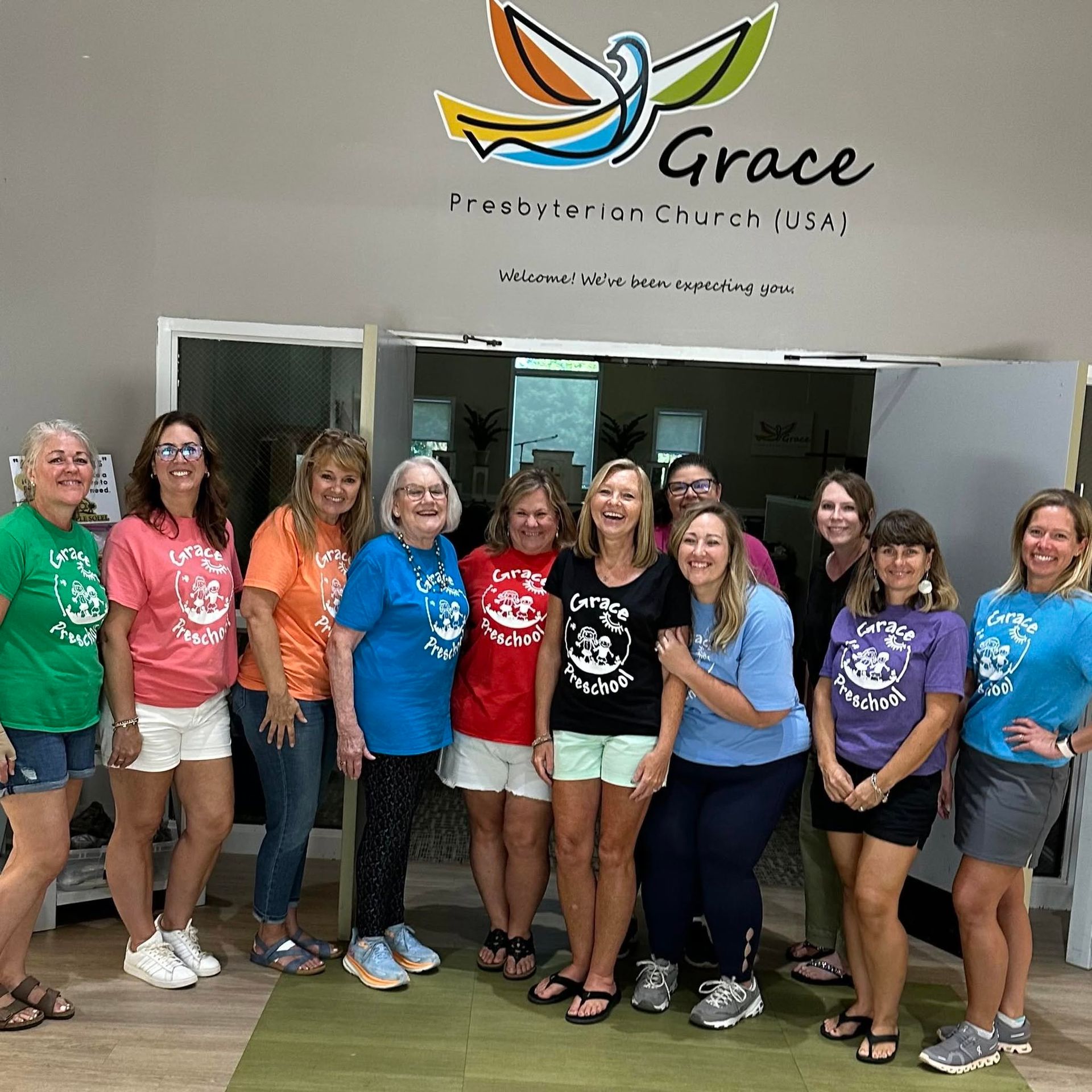 Group of women posing indoors in front of a Grace Fellowship Church sign, smiling in colorful T-shirts.