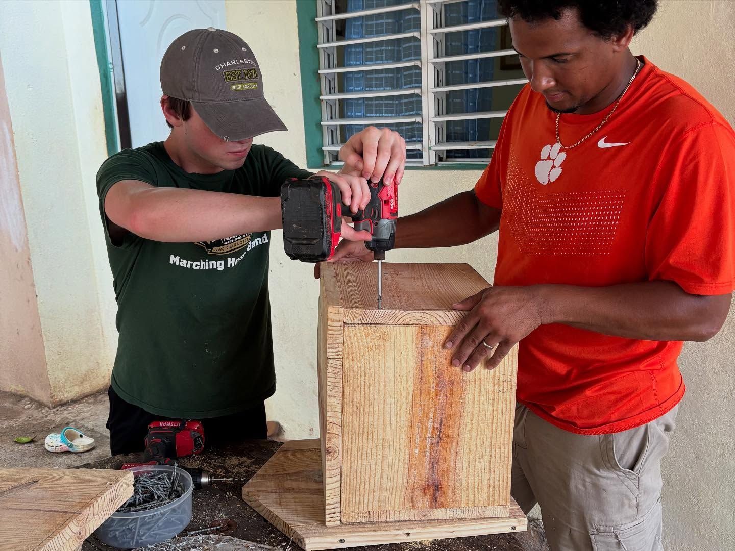 Two people using a power drill to assemble a wooden box outdoors on a porch