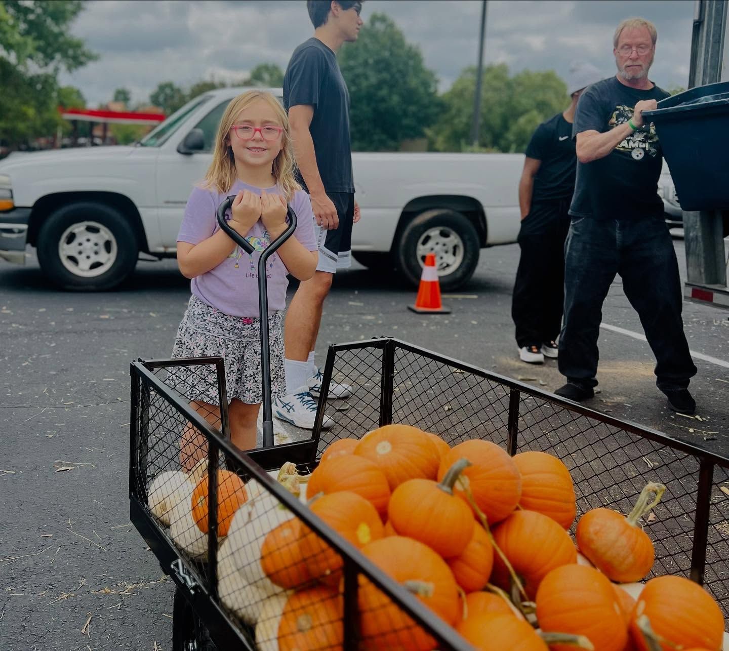 Child holding a pumpkin beside a crate of pumpkins in a parking lot, with adults in the background.