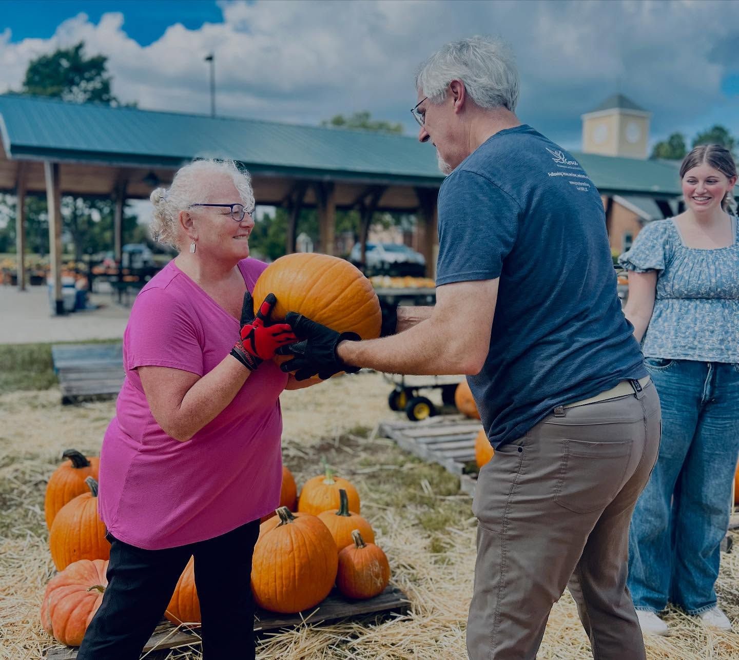 Two people hand a pumpkin at a pumpkin patch, with more pumpkins on the ground.
