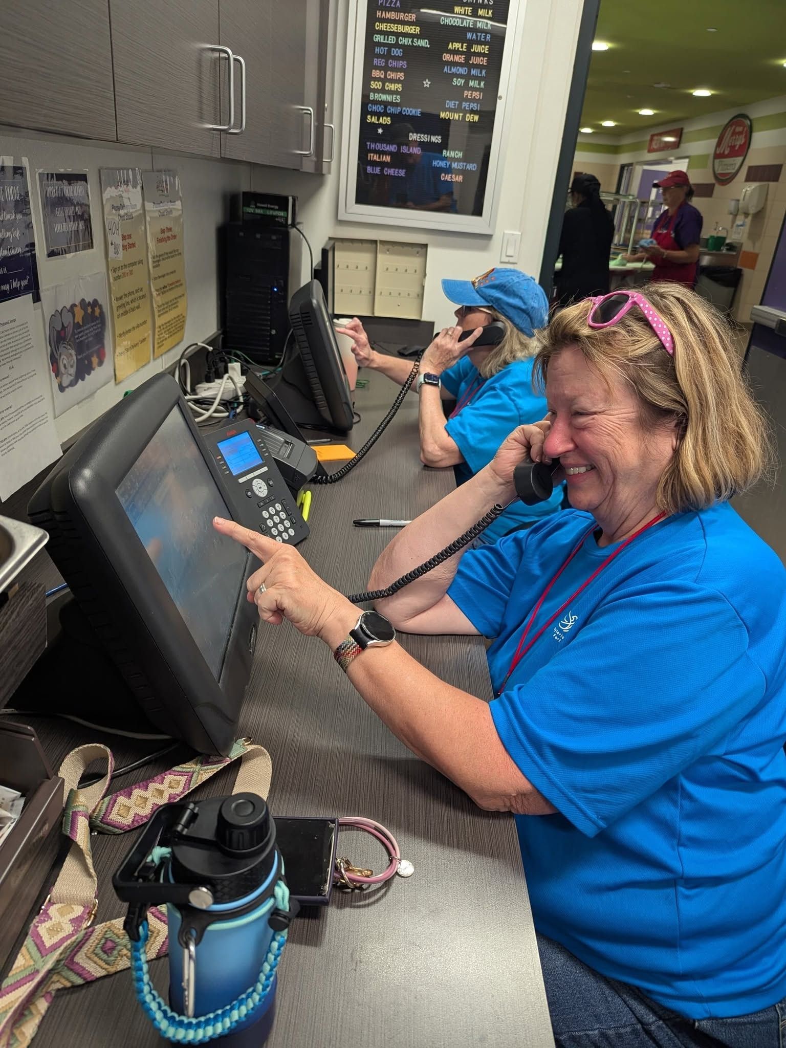 Two people in blue shirts using touchscreens at a counter in a bright indoor workspace