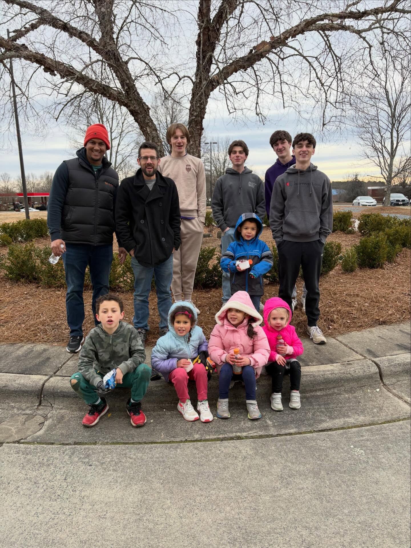 Group of people posing outdoors in a park, with children seated in front and trees in the background.