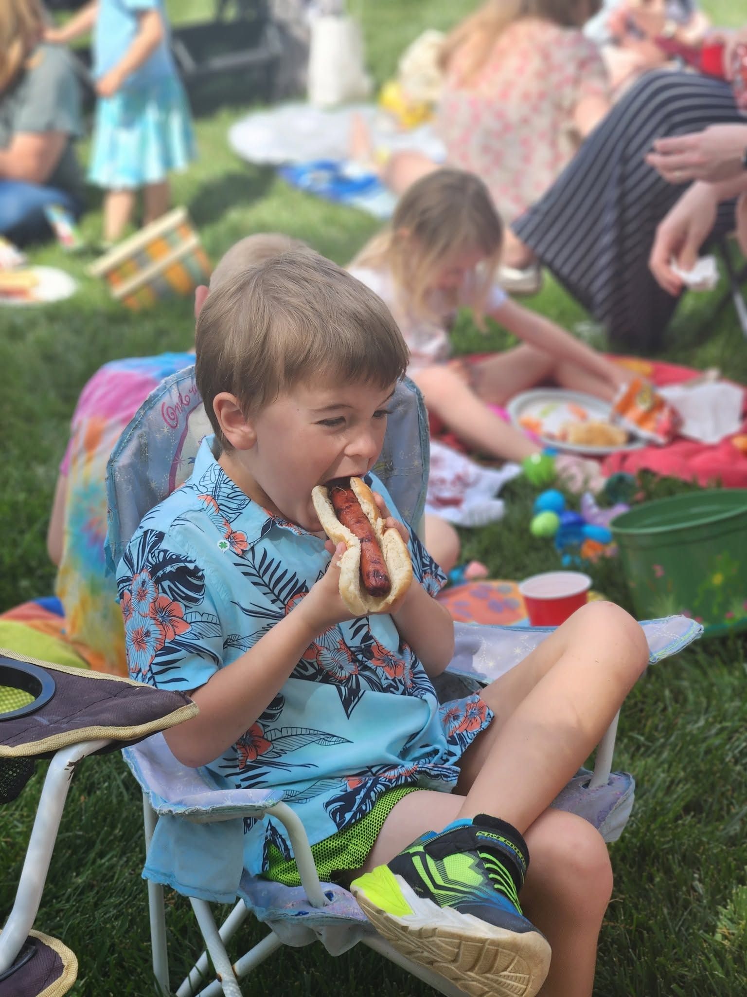Child eating a hot dog while sitting in a lawn chair at an outdoor picnic
