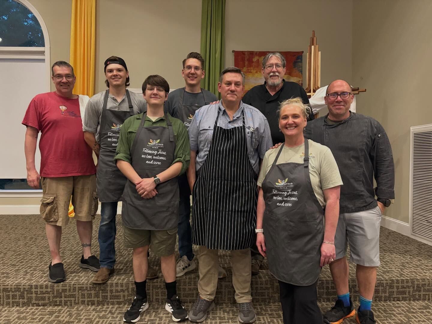 Eight people standing together indoors, some wearing aprons, posing in front of a wall and curtained windows.
