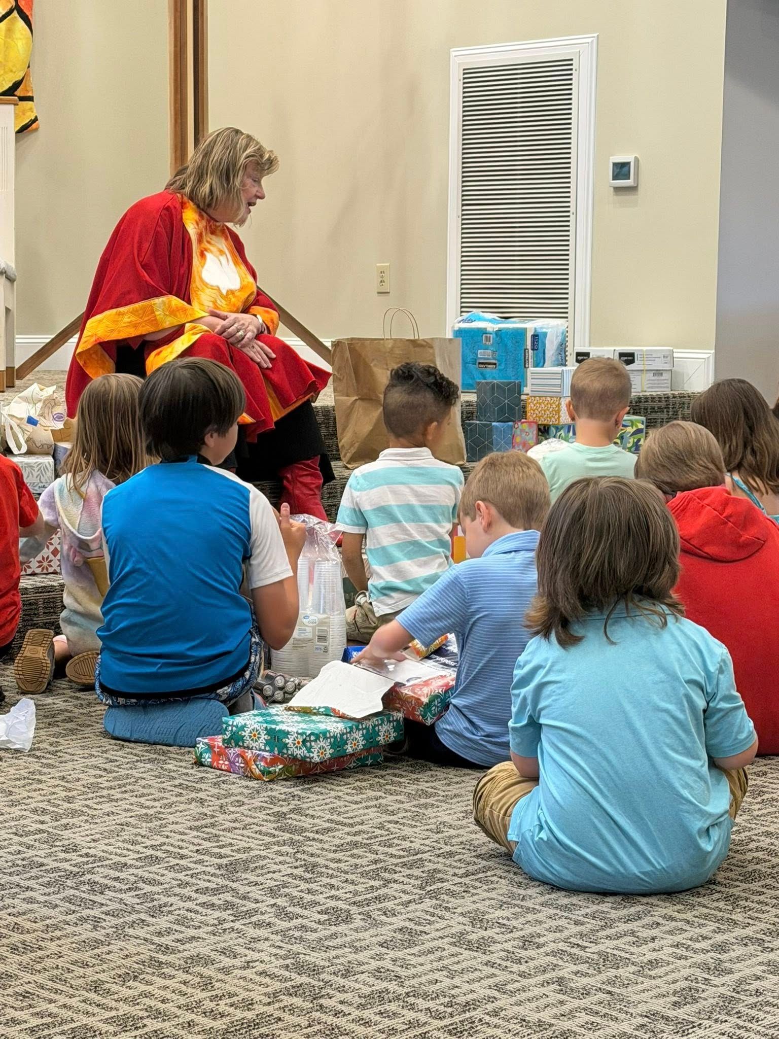 Presenter in red robe sits before a group of children in a carpeted room.