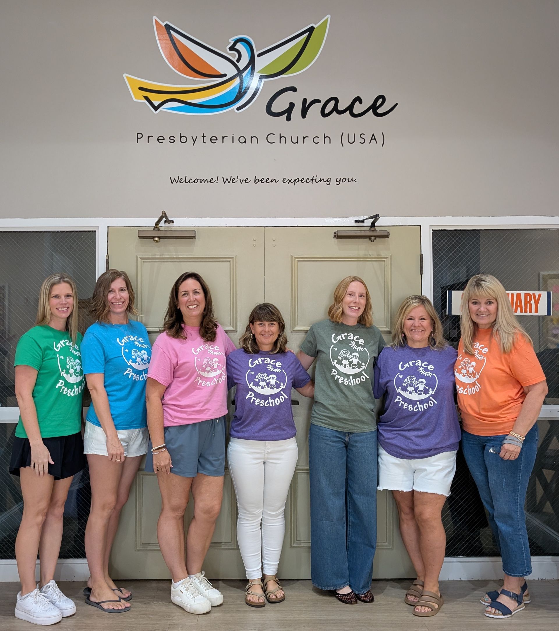 Group of women posing indoors in front of a Grace Fellowship Church sign, smiling in colorful T-shirts.