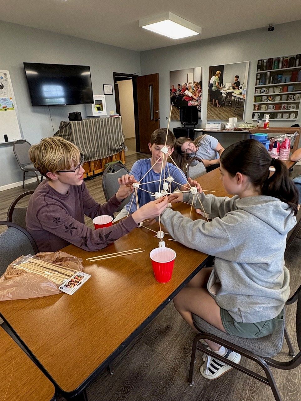 Children at a table making crafts in a classroom, with red cups and papers spread out.