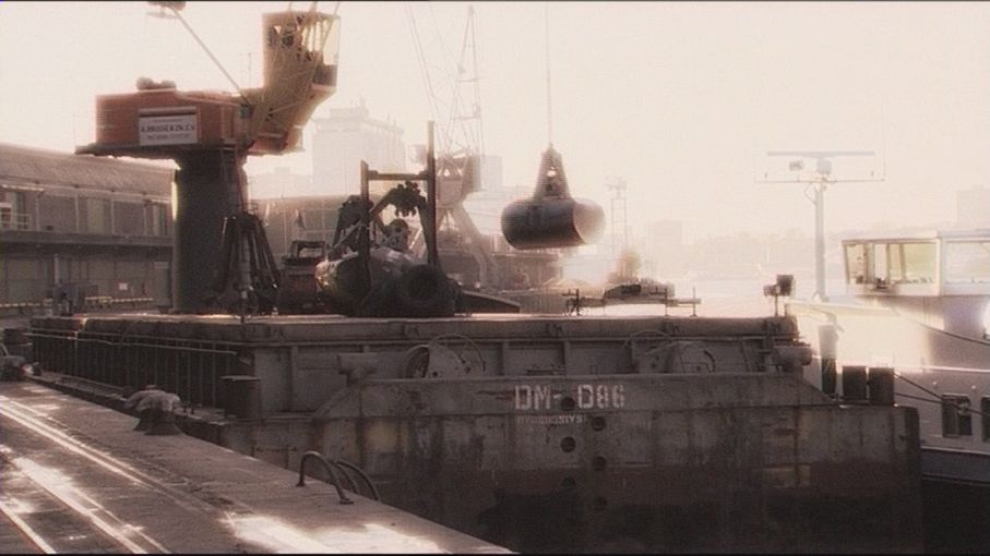 A military ship deck at sunset with cranes, equipment, and smoke rising in the background.