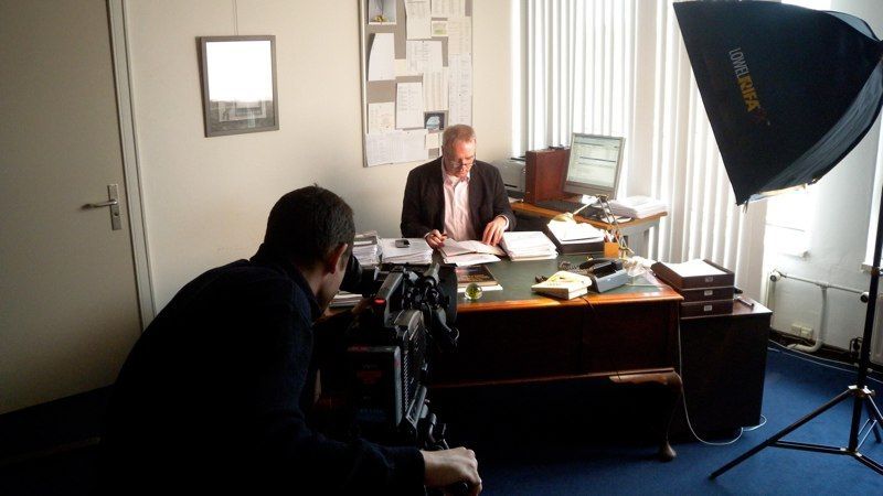 Photographer filming a man at a desk in an office, with studio lighting and a computer nearby