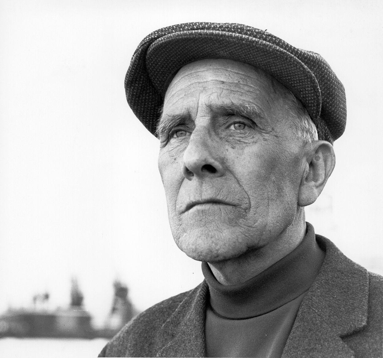 Black-and-white portrait of a man in a flat cap, gazing off to the side outdoors.