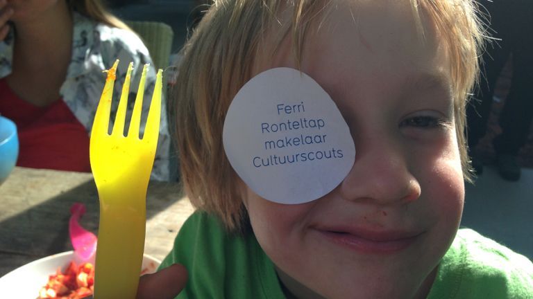 Child smiling with a paper circle over one eye, holding a yellow fork outdoors.