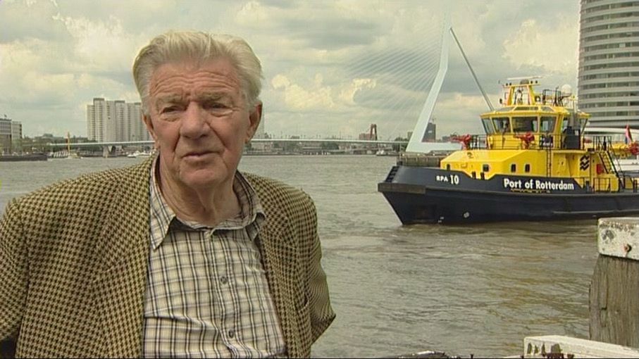 Man standing by a waterfront with a yellow and blue boat in the background