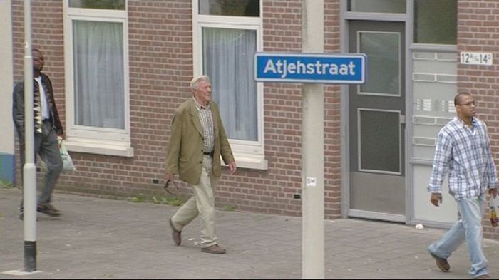 People walking past a brick building and a blue street sign reading “Atjenstraat.”