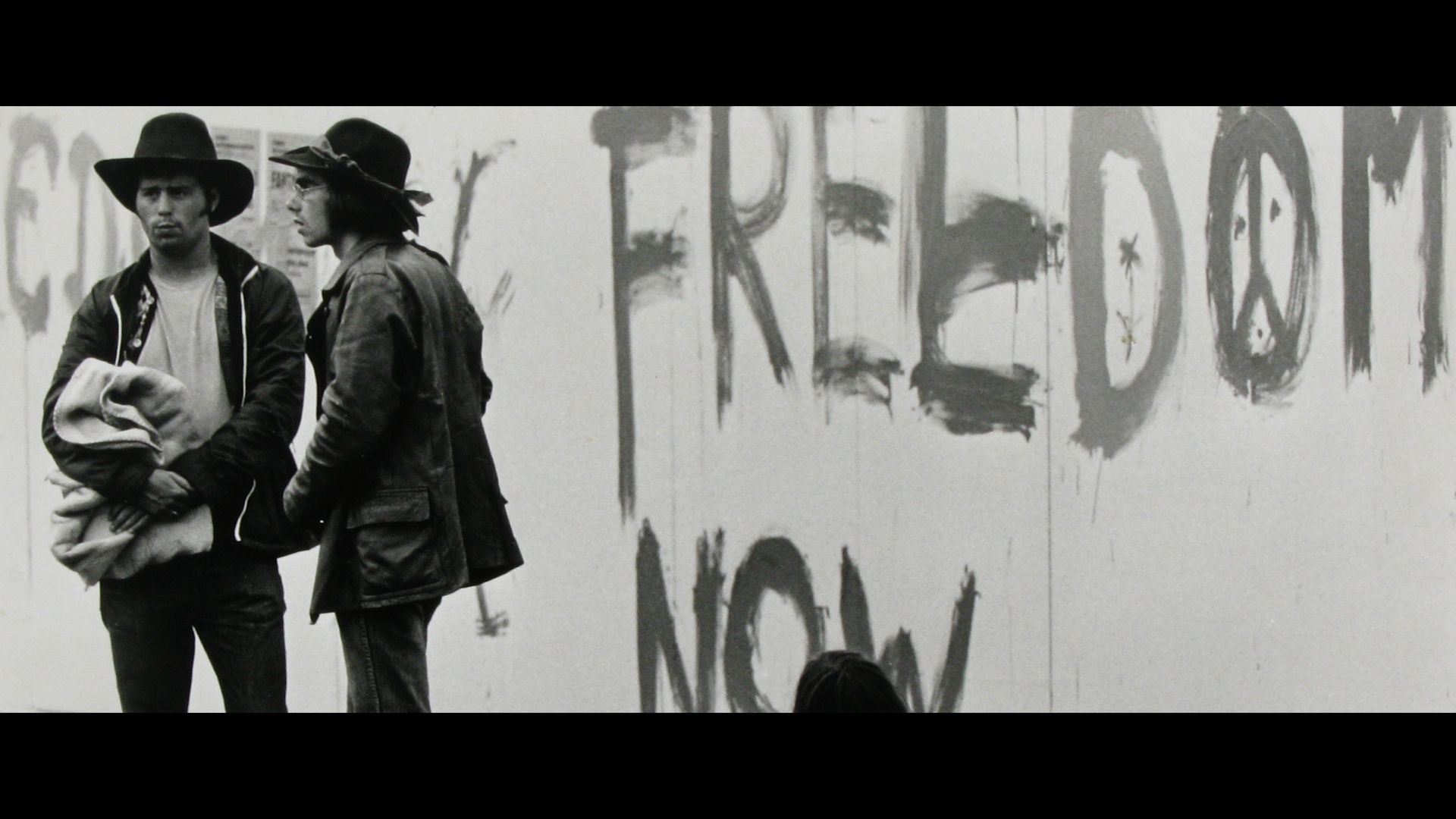 Two people standing by a wall with graffiti reading “FREEDOM NOW”