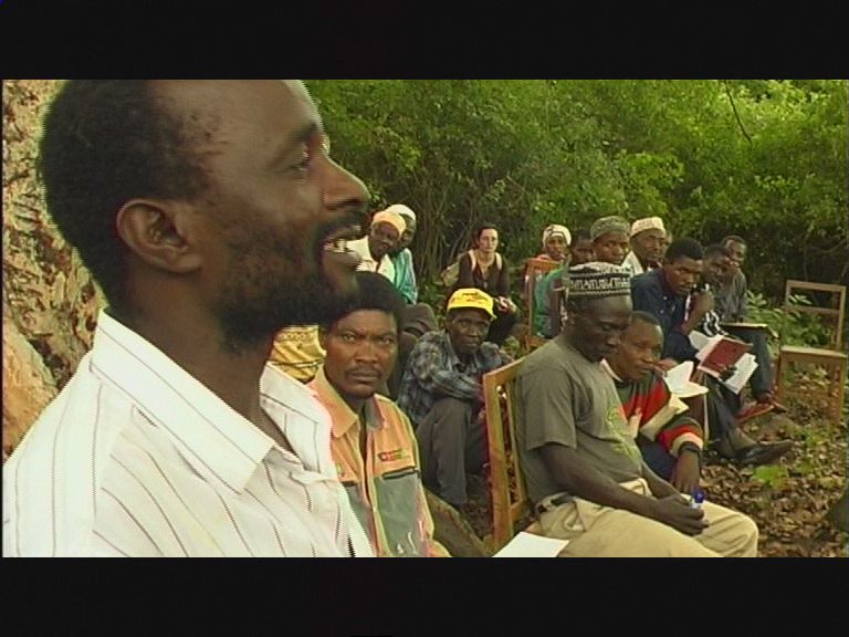 Man speaking to a seated group outdoors near trees, with several people listening attentively.