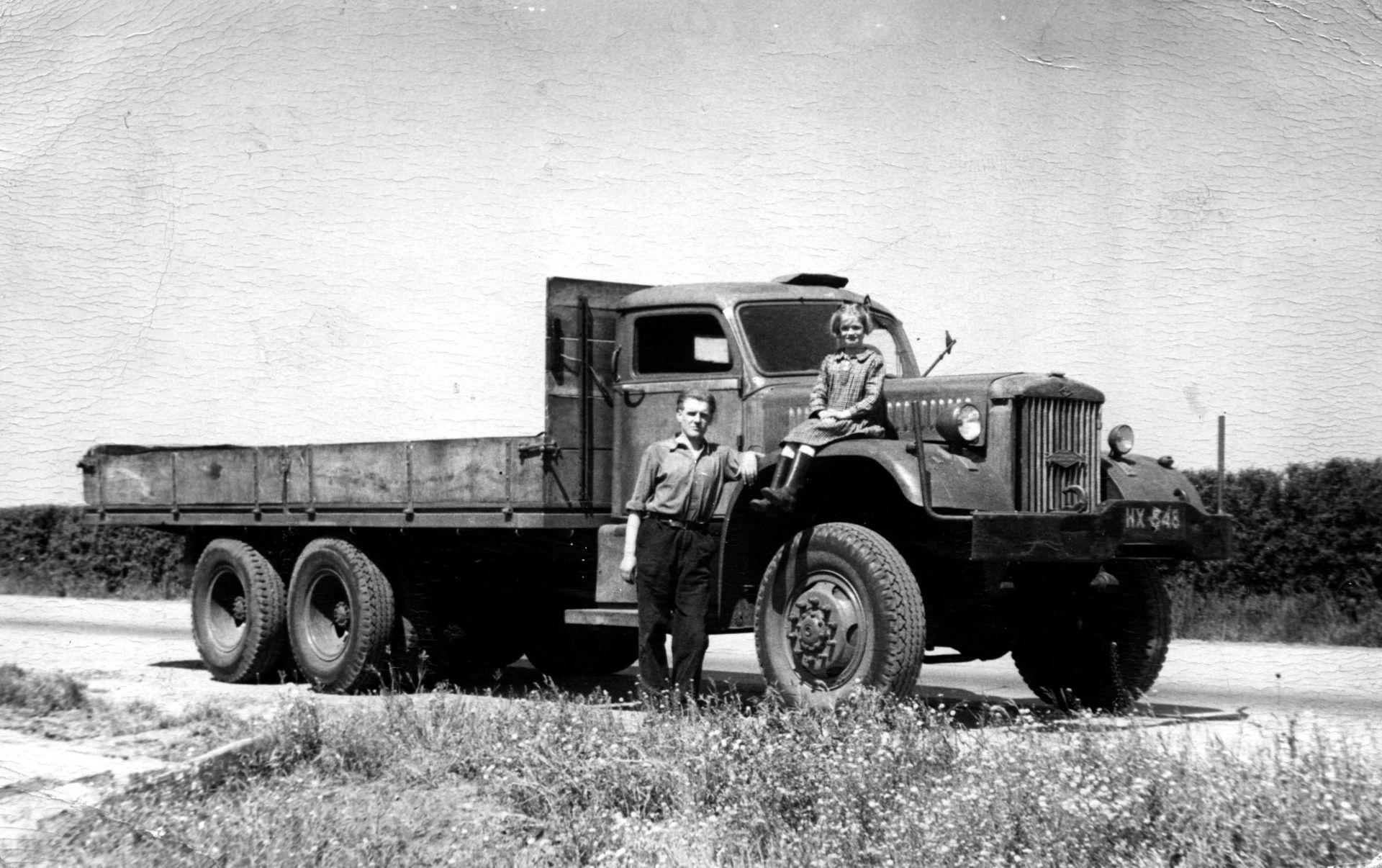 Two men stand beside a vintage flatbed truck on a dirt road in a rural landscape.