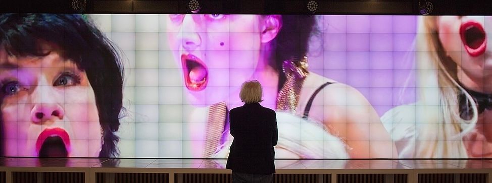Silhouetted person facing a wall of large screens showing close-up faces with open mouths in pink and purple light