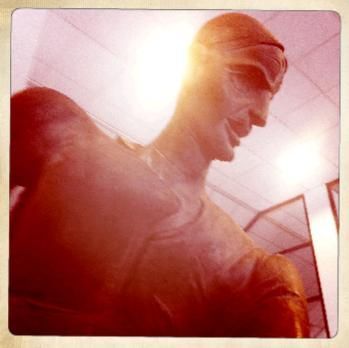 Backlit statue in a museum, viewed from below against a bright ceiling.