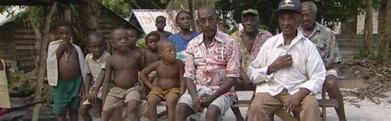 A group of people seated outdoors under a tree in a rural setting, facing the camera.