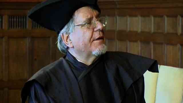 Person in black academic regalia and cap holding a yellow folder, standing indoors before wood paneling.