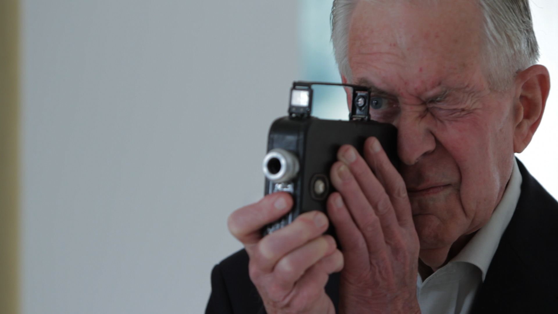 Man using a vintage camera to his eye, aiming it in a plain indoor setting