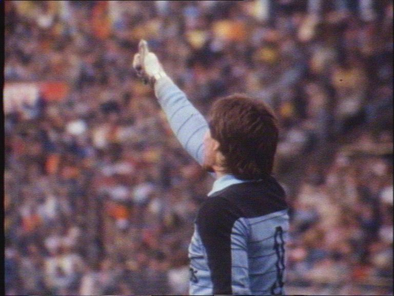 Athlete in dark jersey raises an arm toward a cheering stadium crowd