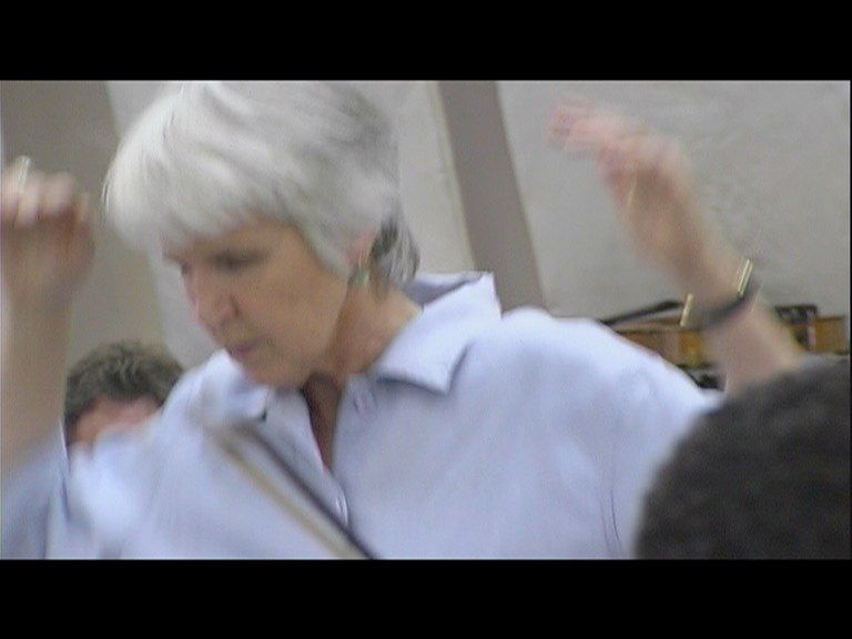 Person with short white hair in a light blue shirt raising their arms indoors