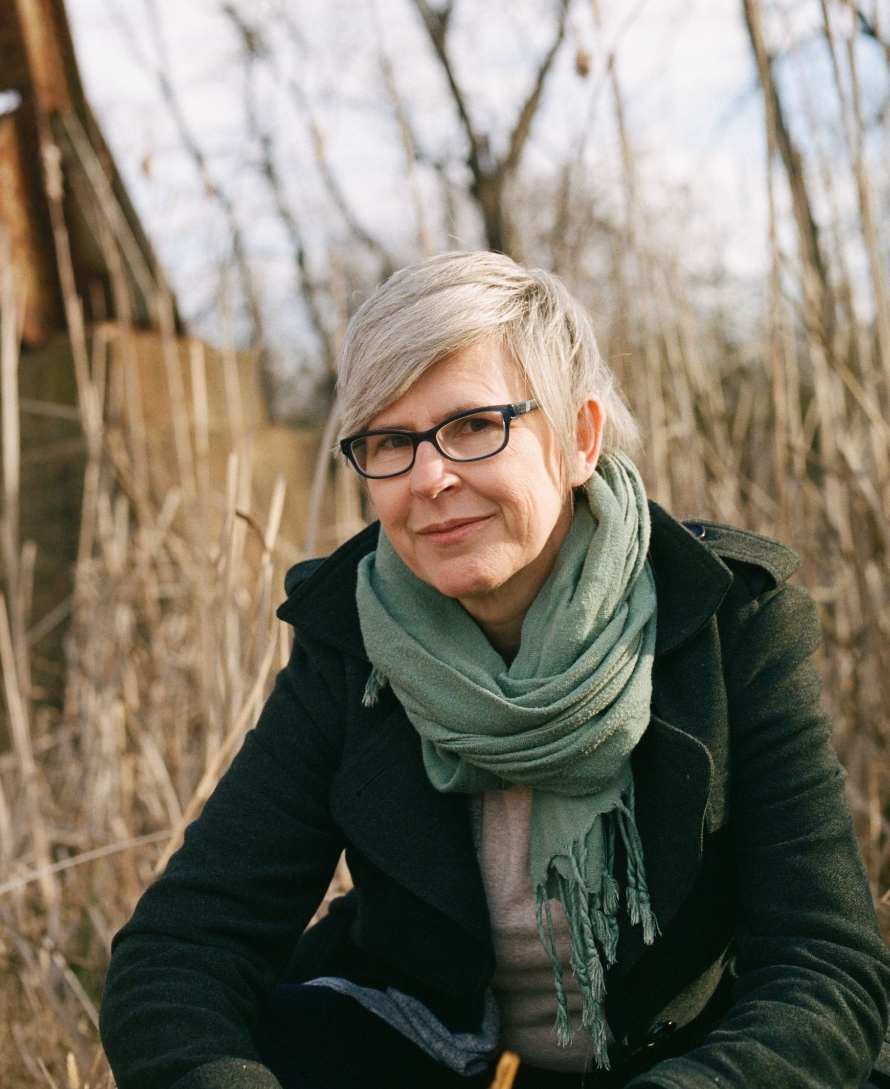 picture of Lisa Kammerud LMT, grey hair, glasses, smiling slightly, in front of a field of brown grasses