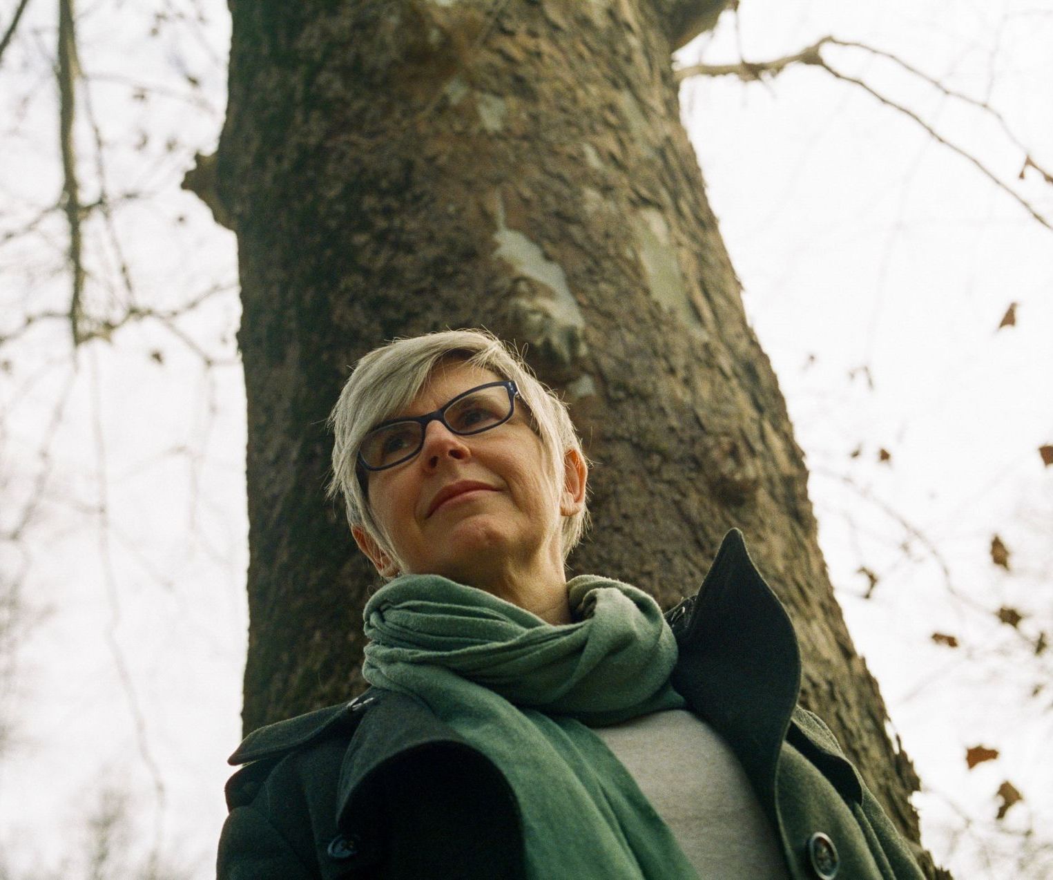 LIsa Kammerud LMT, wearing glasses and a green scarf, standing in front of a tree. They embody their wisdom and power