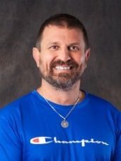 A man with a beard is wearing a blue champion t-shirt and a necklace.