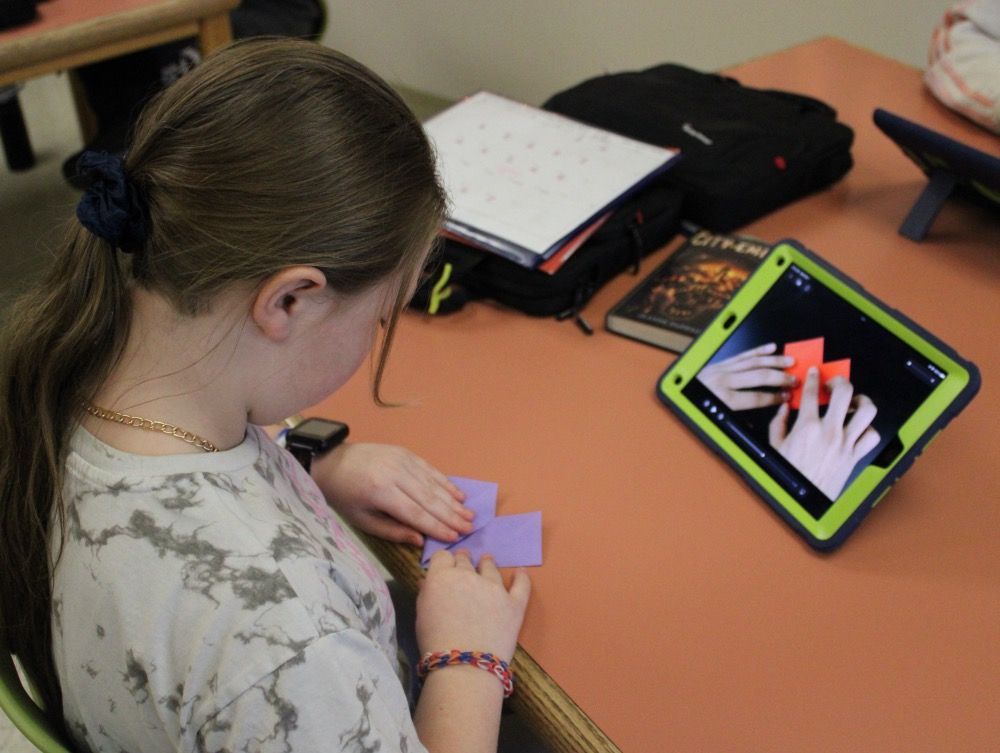A girl sits at a table looking at a tablet with a picture of her hands on it