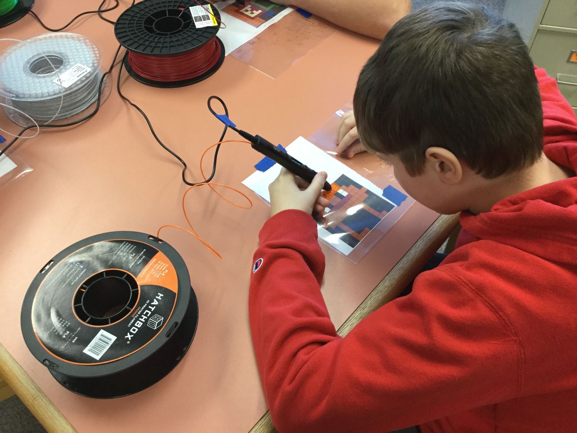 A boy in a red hoodie is using a pen to draw on a piece of paper