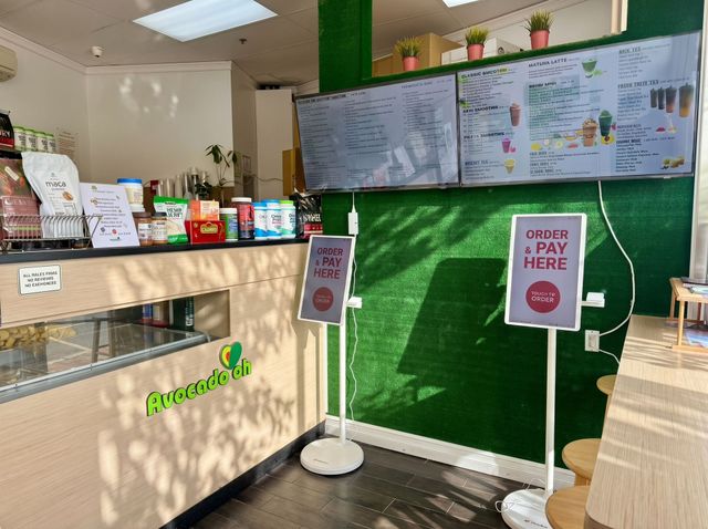 Bright café counter with menu boards, green wall, and “Please Pay Here” signs near the register.