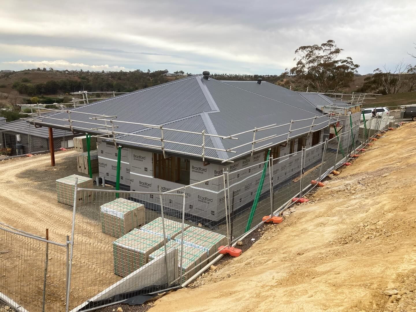 Partially built house with grey roof framing on a sloped dirt lot, surrounded by temporary fencing — Twomey Homes in Delacombe, VIC