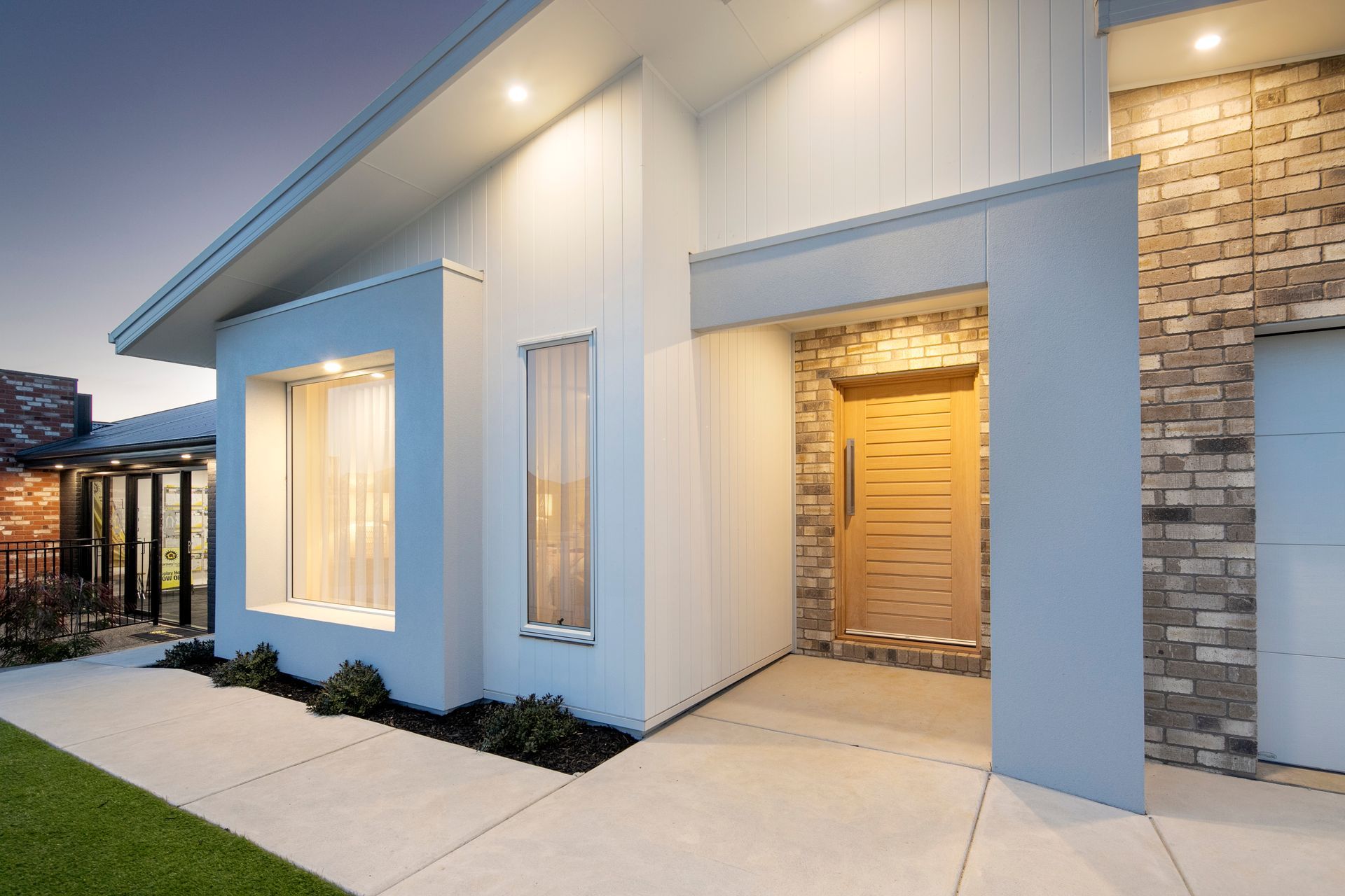 Modern white house exterior with recessed front entry, brick accent wall, and concrete walkway at dusk — Twomey Homes in Delacombe, VIC