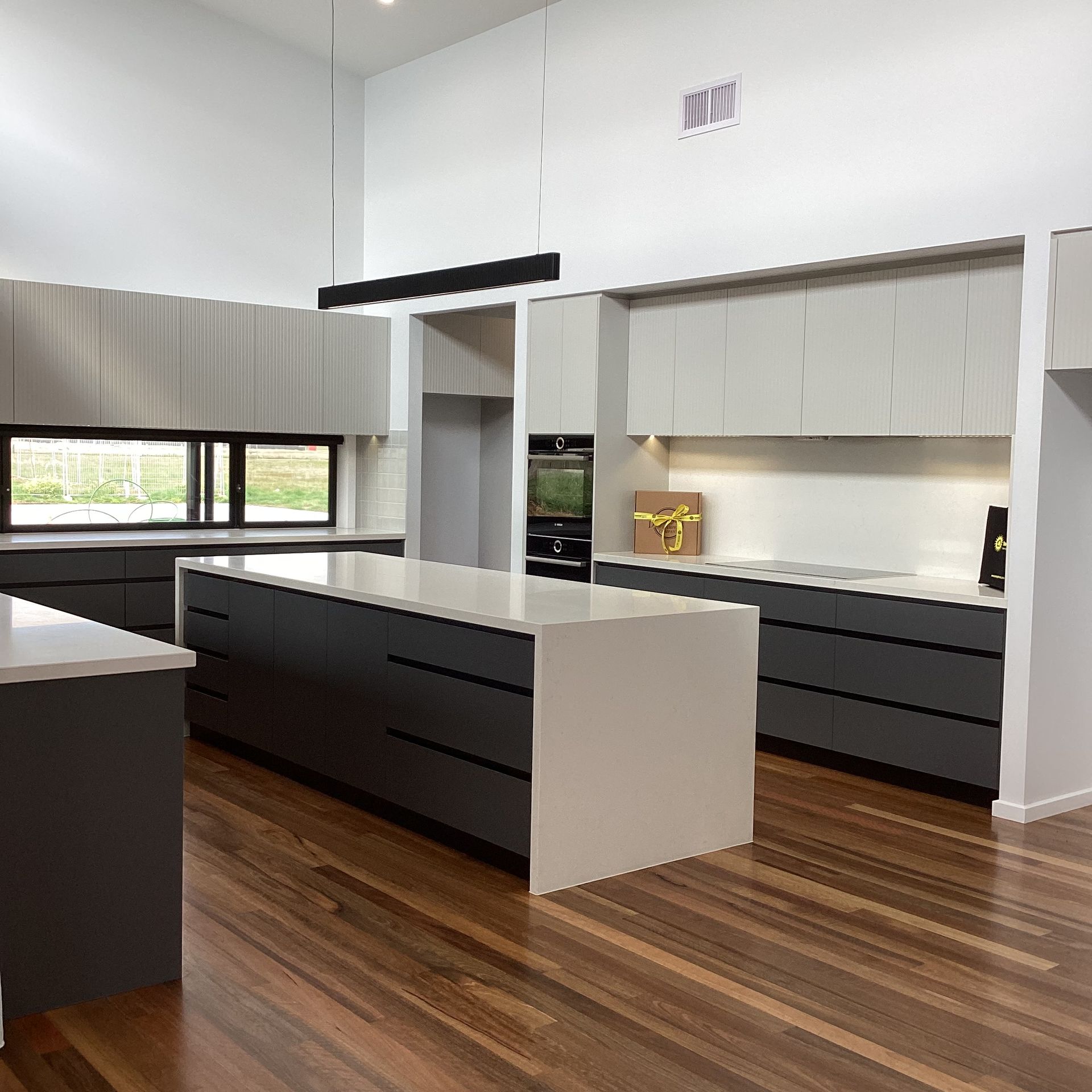 Modern kitchen with white walls, dark grey cabinets, wood floors, and a large island — Twomey Homes in Delacombe, VIC