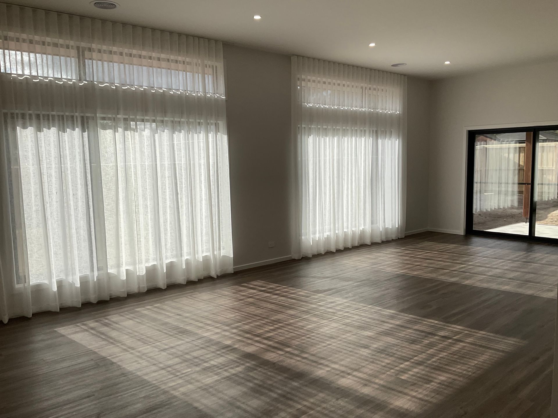 Empty sunlit living room with sheer white curtains, wood floor, and large windows — Twomey Homes in Delacombe, VIC
