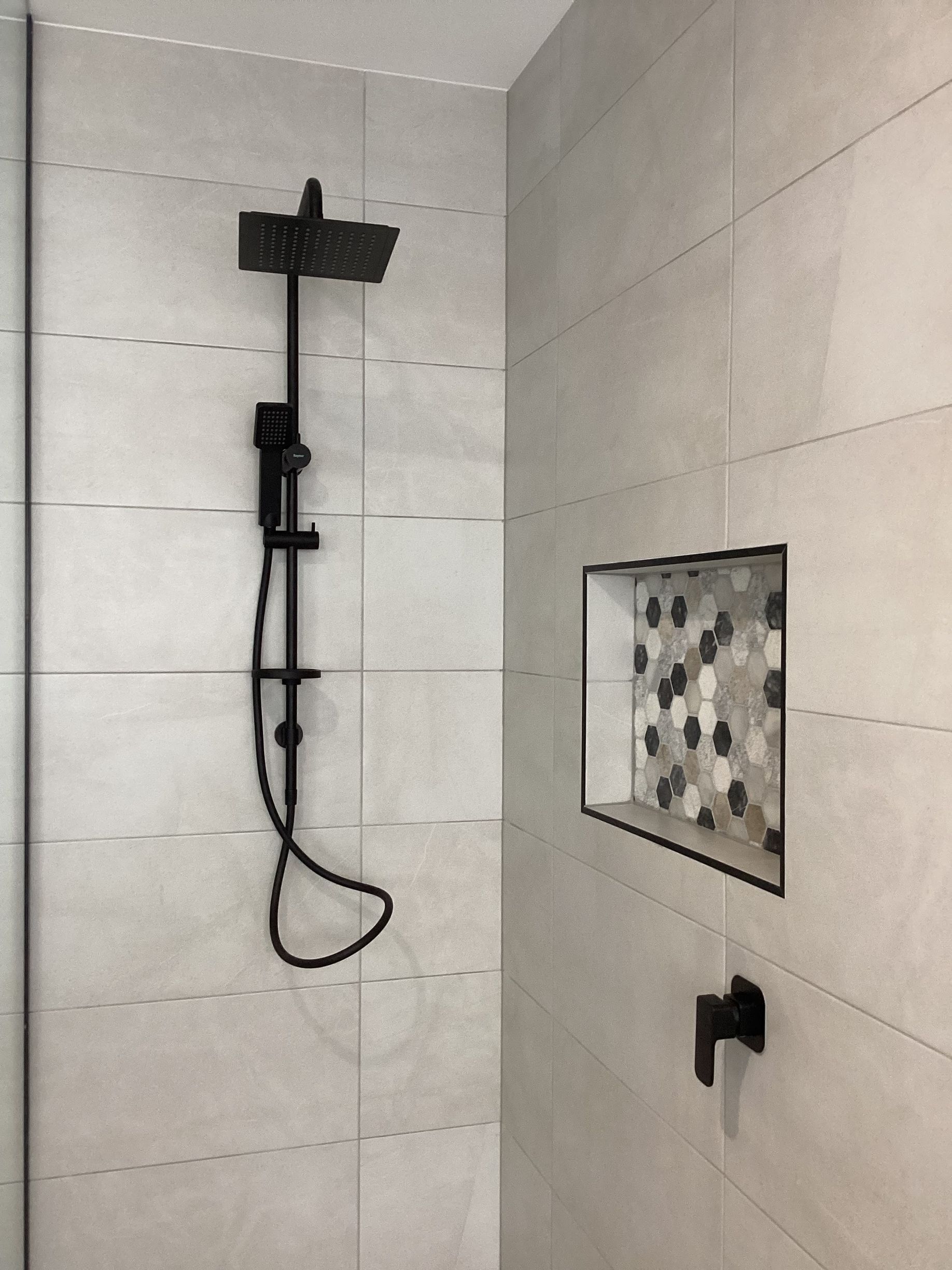Modern tiled shower with black rain showerhead and handheld sprayer, plus a recessed shelf with mosaic tile accents — Twomey Homes in Delacombe, VIC