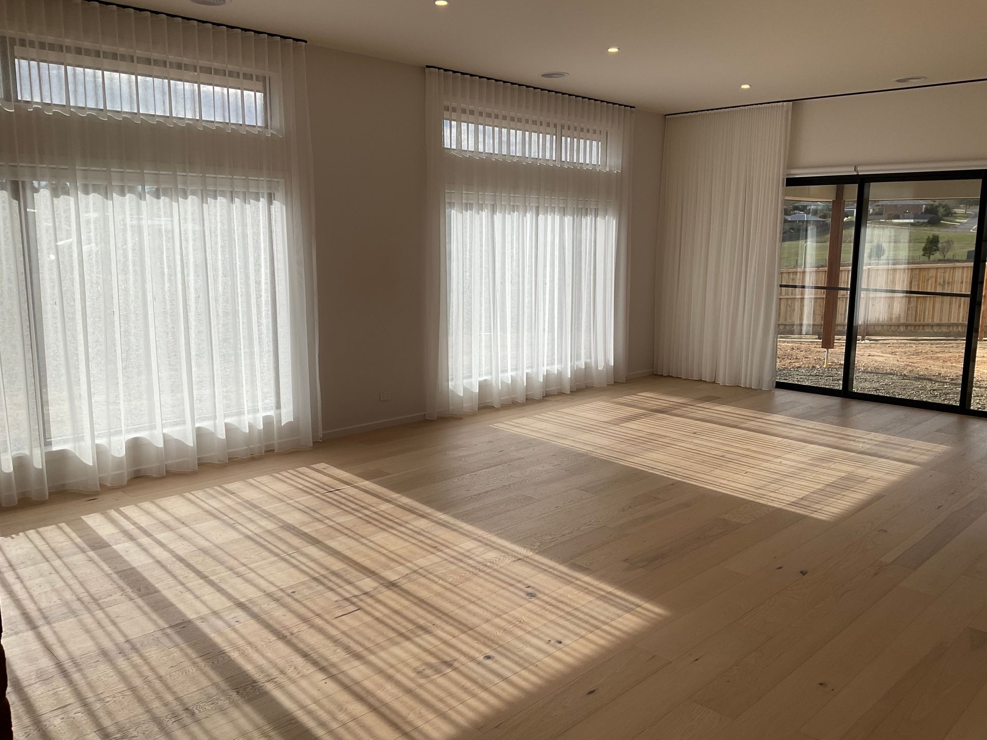 Empty sunlit room with large sheer-curtained windows and sliding glass doors, casting long shadows on the floor — Twomey Homes in Delacombe, VIC