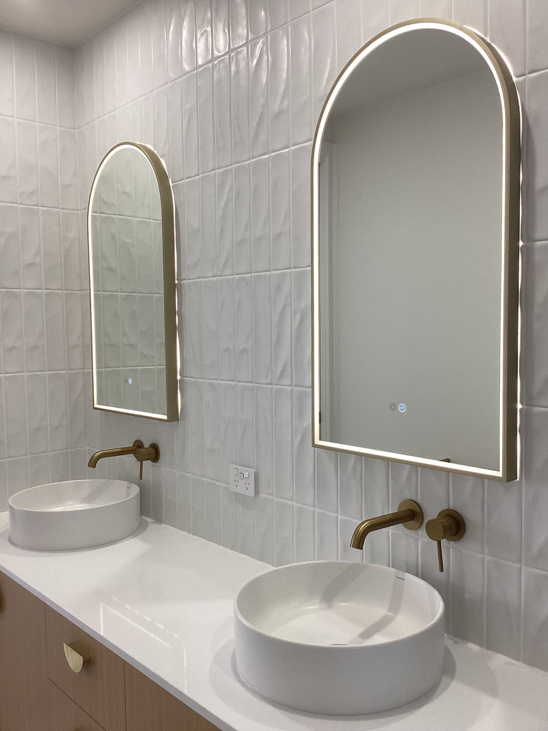 Modern bathroom with two round sinks, brass taps, and arched mirrors on white tiled walls — Twomey Homes in Delacombe, VIC