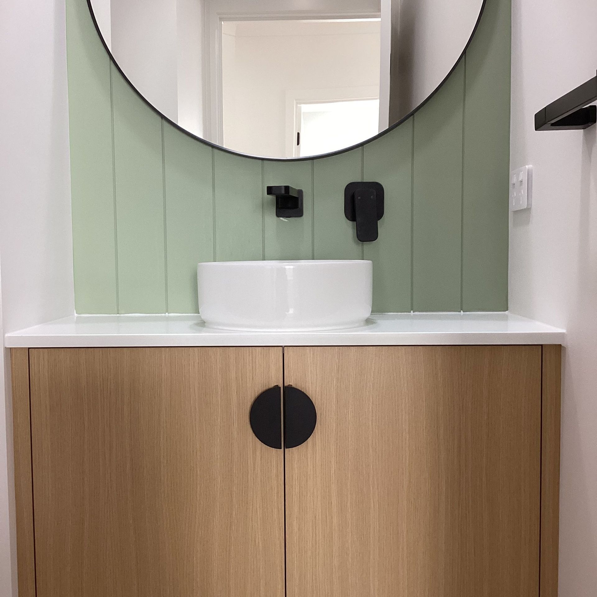 Modern bathroom vanity with round mirror, white sink, and wood cabinet in a green-panelled wall — Twomey Homes in Delacombe, VIC