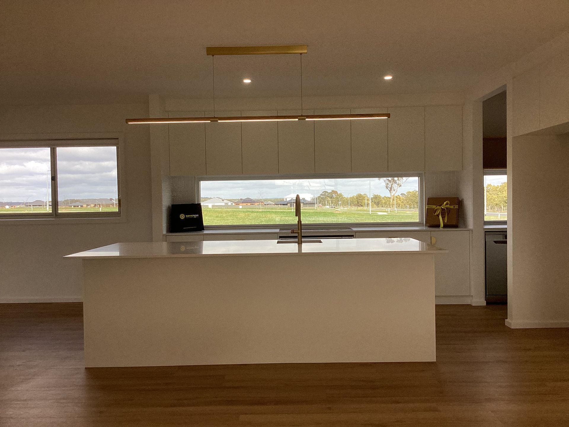 Modern white kitchen with island, sink, wood floors, and large windows overlooking a grassy view — Twomey Homes in Delacombe, VIC