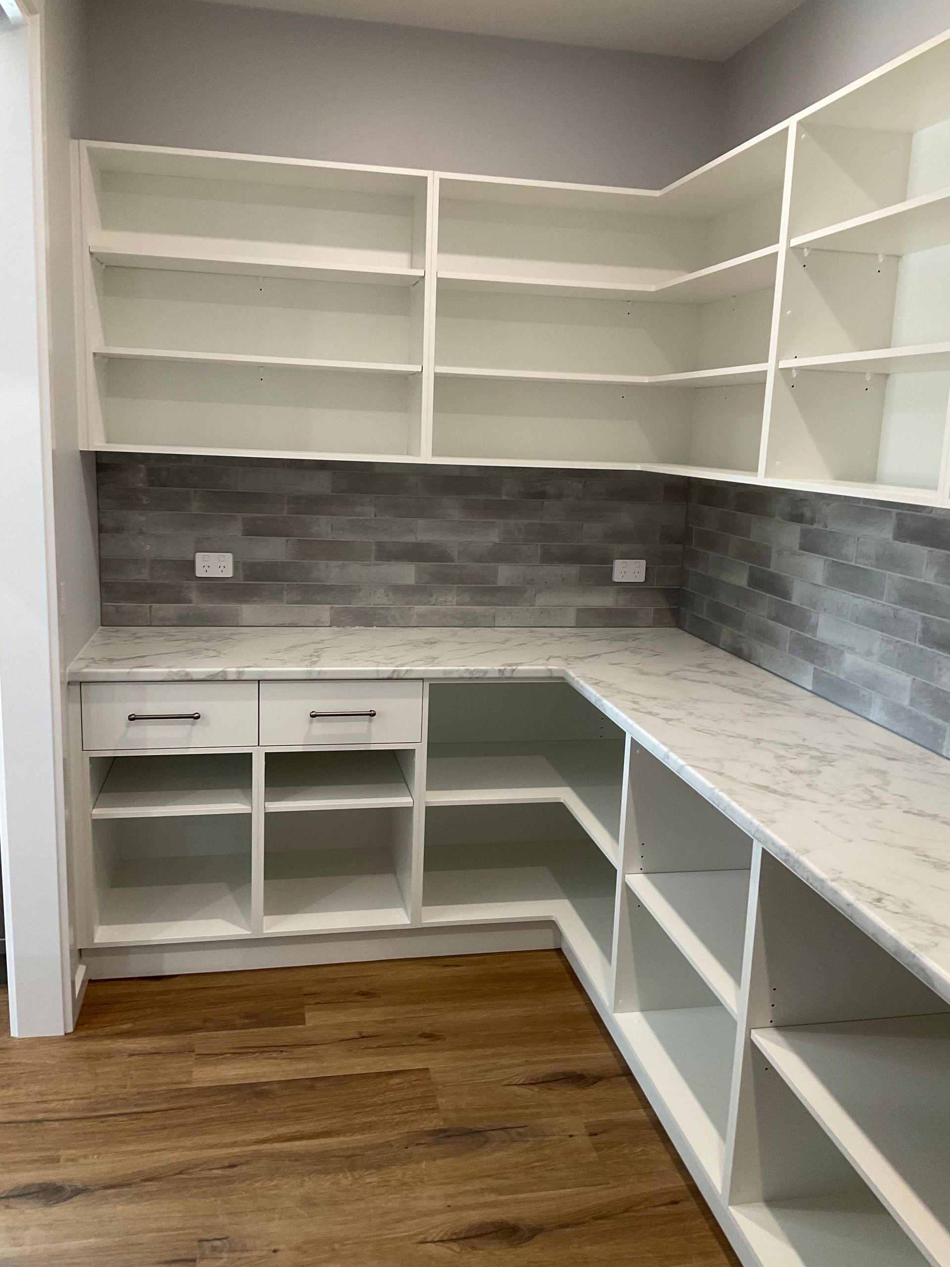 Empty L-shaped kitchen with white shelves, grey tile backsplash, and unfinished wood floor — Twomey Homes in Delacombe, VIC