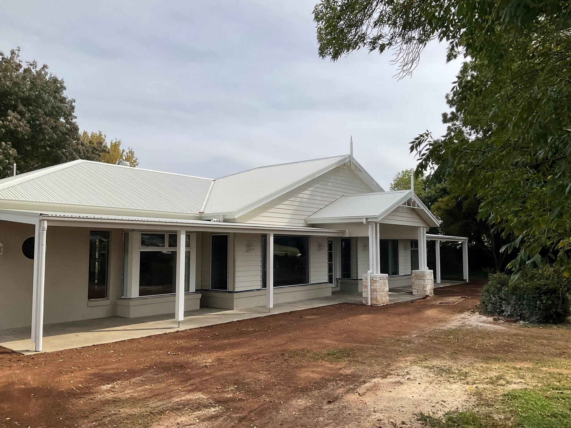 White farmhouse with wraparound porch and red dirt yard under cloudy sky — Twomey Homes in Delacombe, VIC