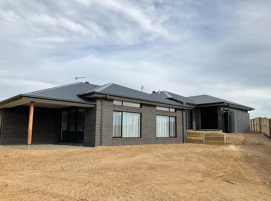 Modern single-story brick house with a dark roof on a bare dirt lot under a cloudy sky — Twomey Homes in Delacombe, VIC