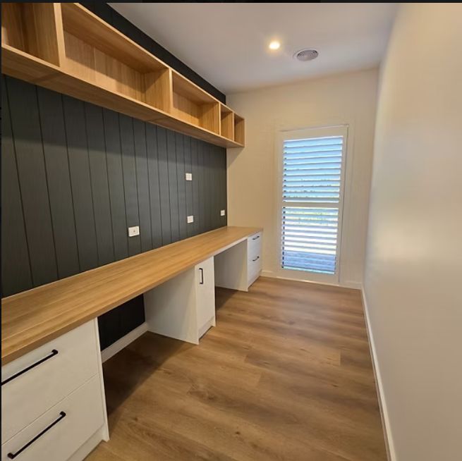 Small modern home office with wood desk, dark panelled wall, white cabinets, and a window with blinds — Twomey Homes in Delacombe, VIC