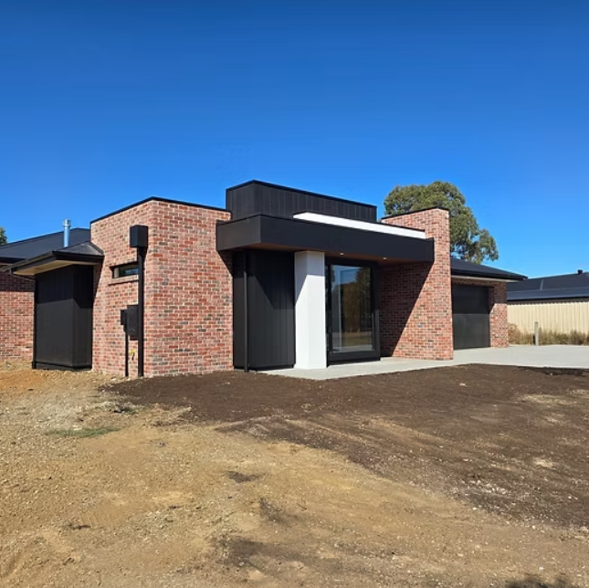 Small modern brick house with a dark awning on a dirt lot under a clear blue sky — Twomey Homes in Delacombe, VIC