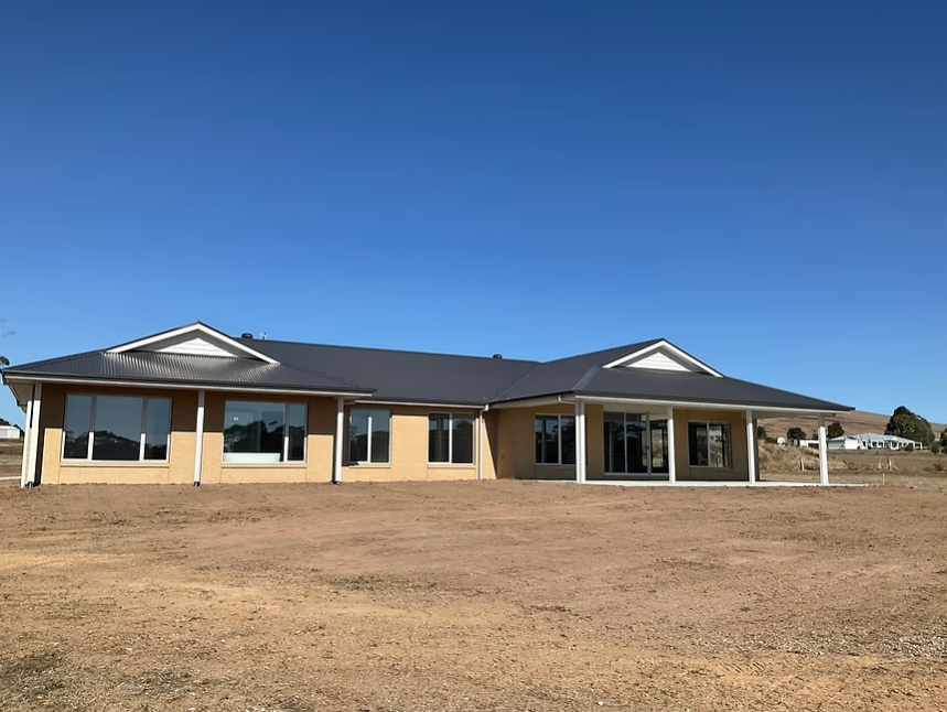Single-story tan house with dark roof on a bare lot under a clear blue sky — Twomey Homes in Delacombe, VIC