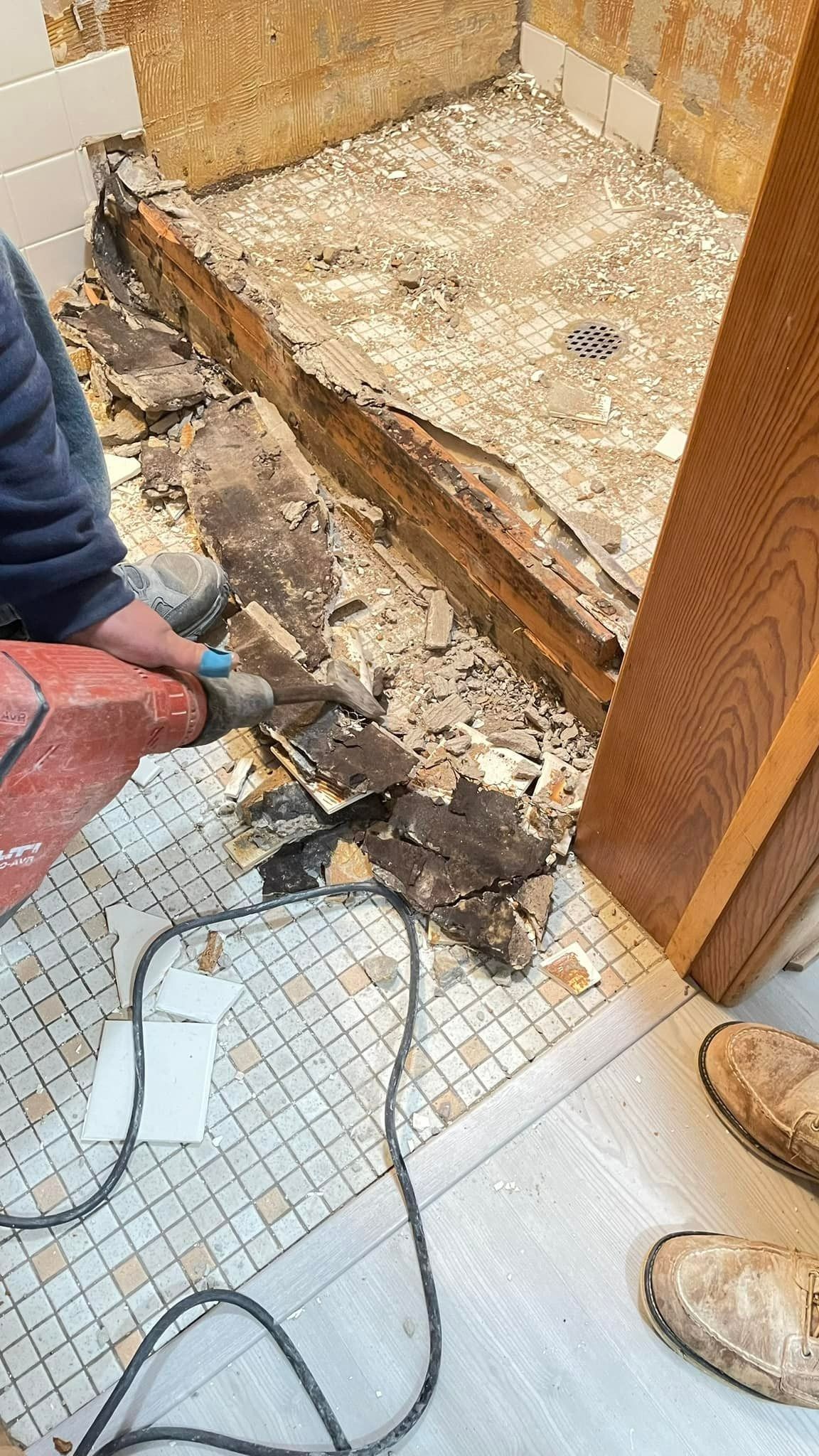 Person using a jackhammer to remove damaged shower flooring. Demolition in progress with debris visible.