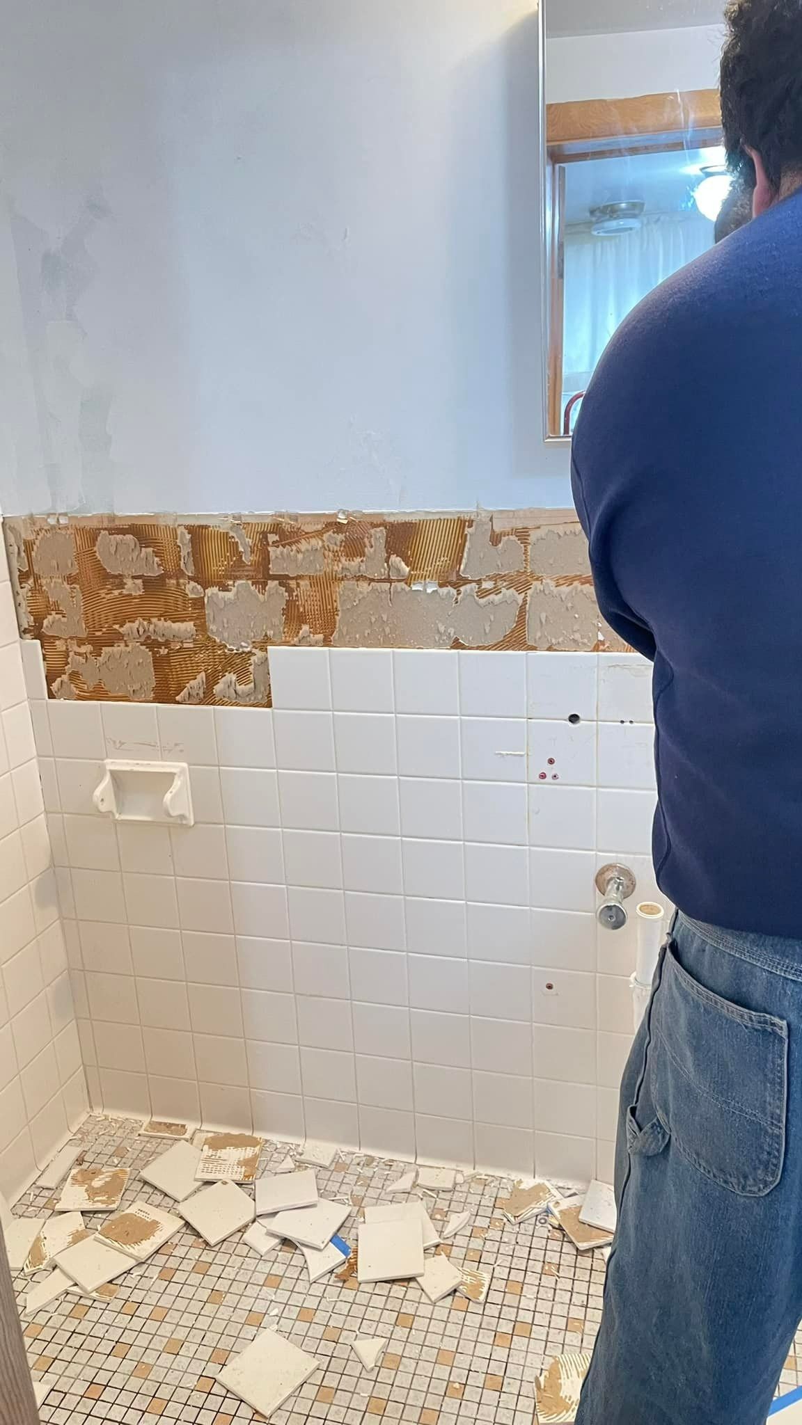 A person stands in a bathroom undergoing renovation, showing a partially tiled wall with loose debris on the floor.